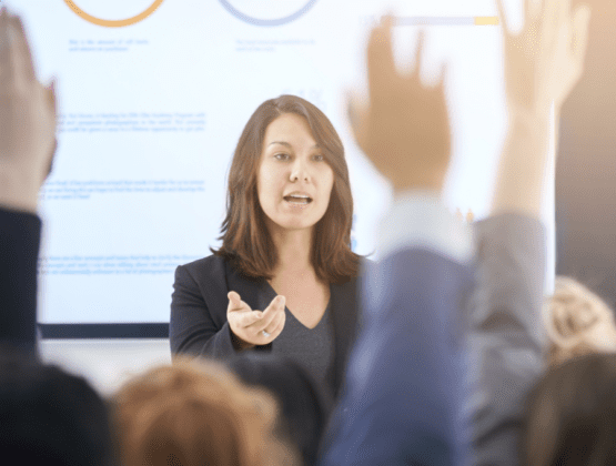 Woman leading a presentation. Some people in the audience have their hands up.