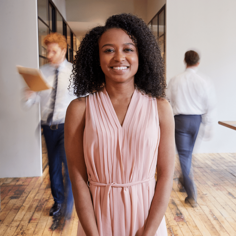 Female office employee stood smiling in a busy office environment.