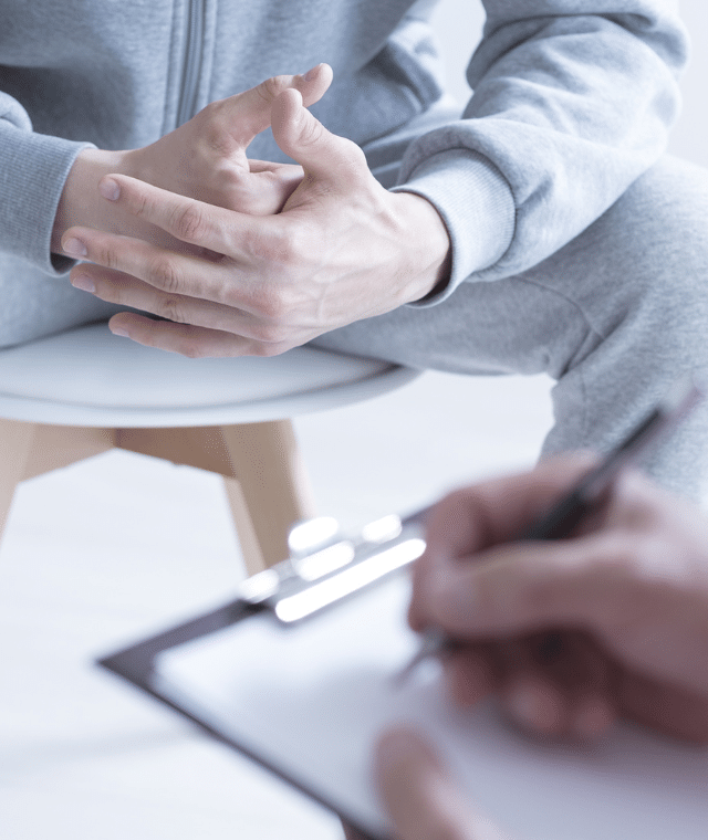 Two people speaking in a clinical environment. One is wearing joggers and the other has a clipboard and is writing notes.