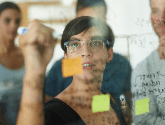 Woman writing on a glass wall with whiteboard pen, colleagues stood behind her.