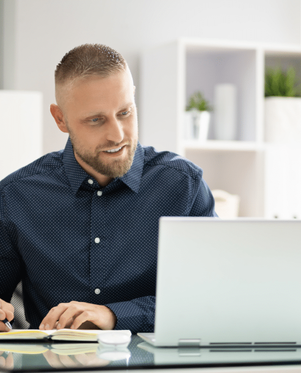 Man working on laptop and taking notes in a notebook with a pen.