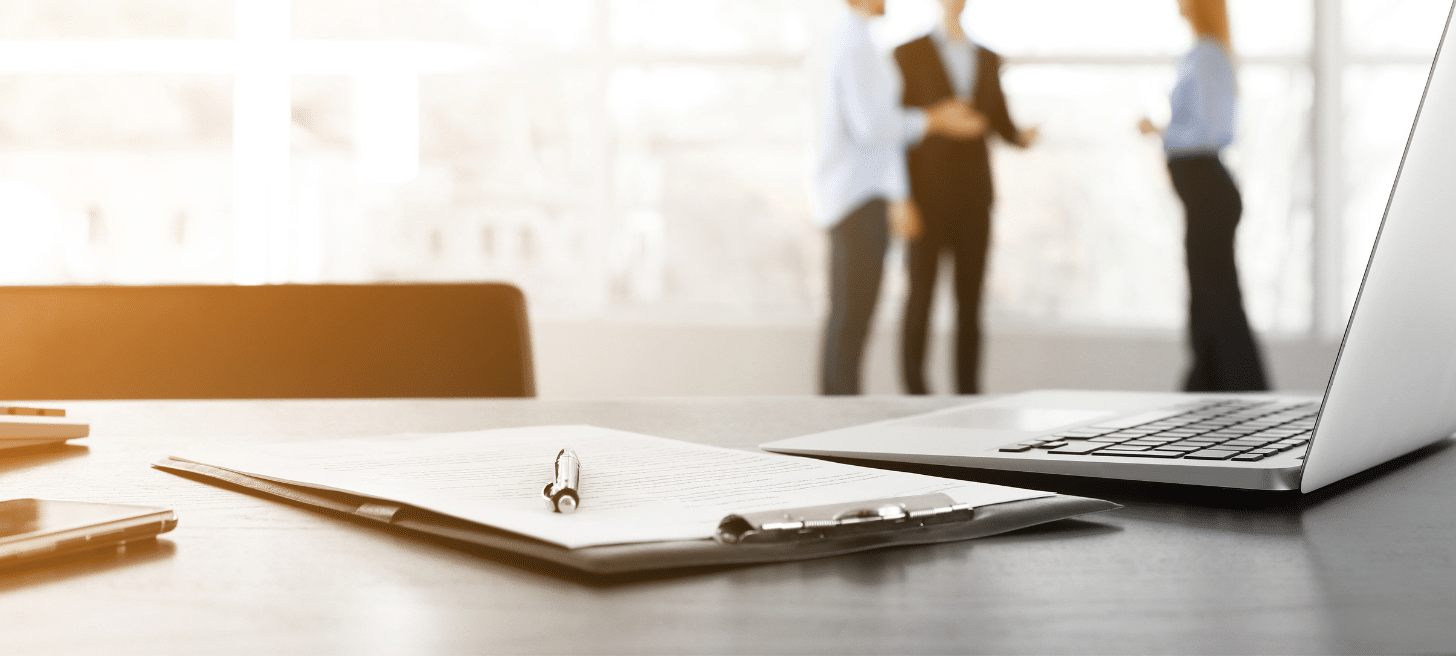 A close-up of a pen on a table in an office with three office workers in the background