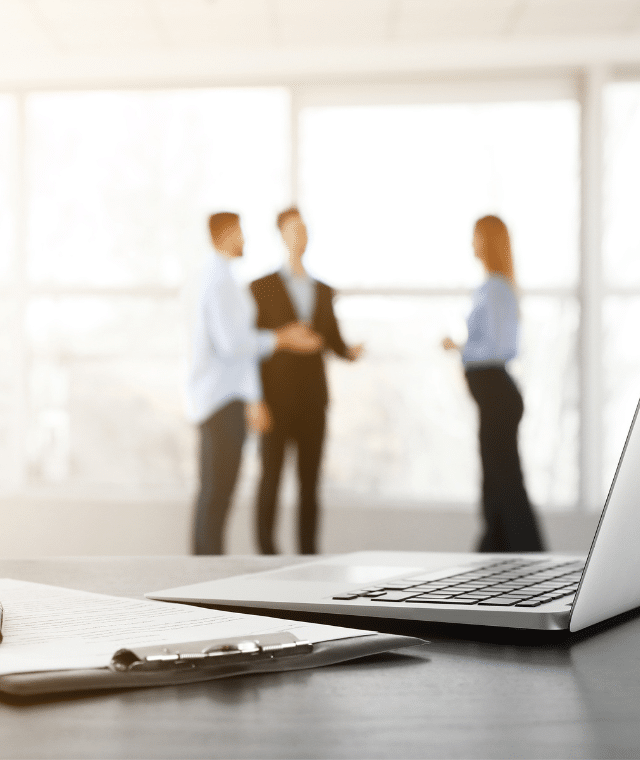 A close-up of a pen on a table in an office with three office workers in the background