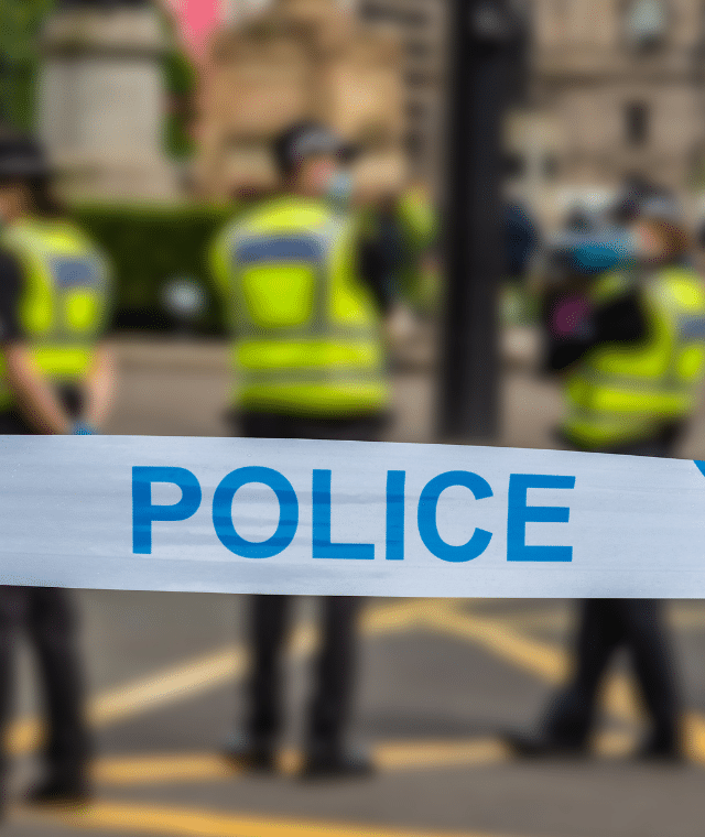 Police tape across public space. Three police officers stood in background.