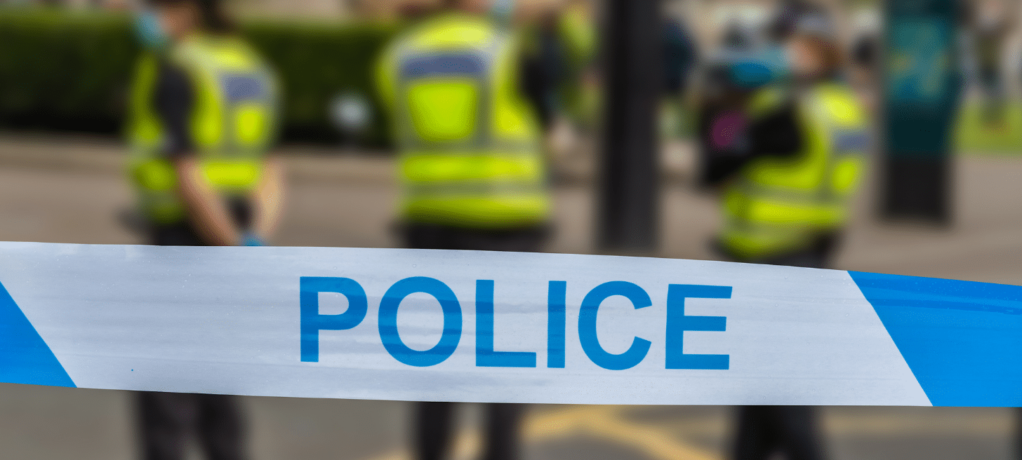 Police tape across public space. Three police officers stood in background.