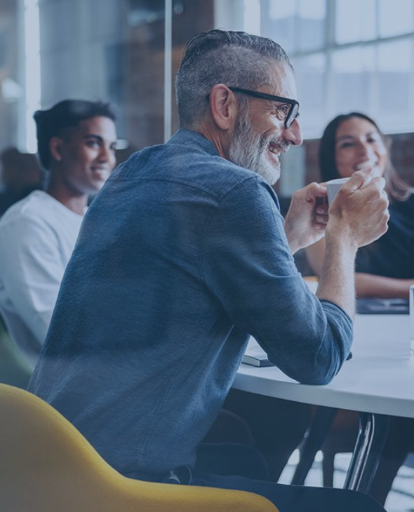 A white haired man with a bear drinks a coffee in a meeting with colleagues.