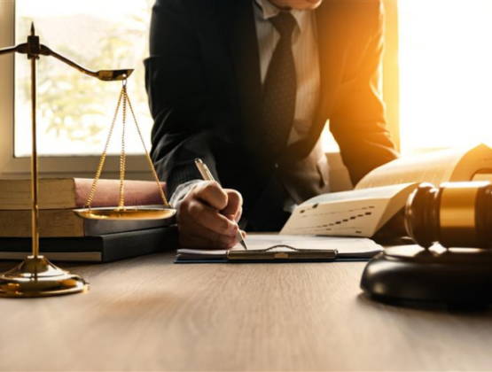 A person in a suit writes on a clipboard at a desk with legal scales, books, and a gavel, illuminated by sunlight from a window.